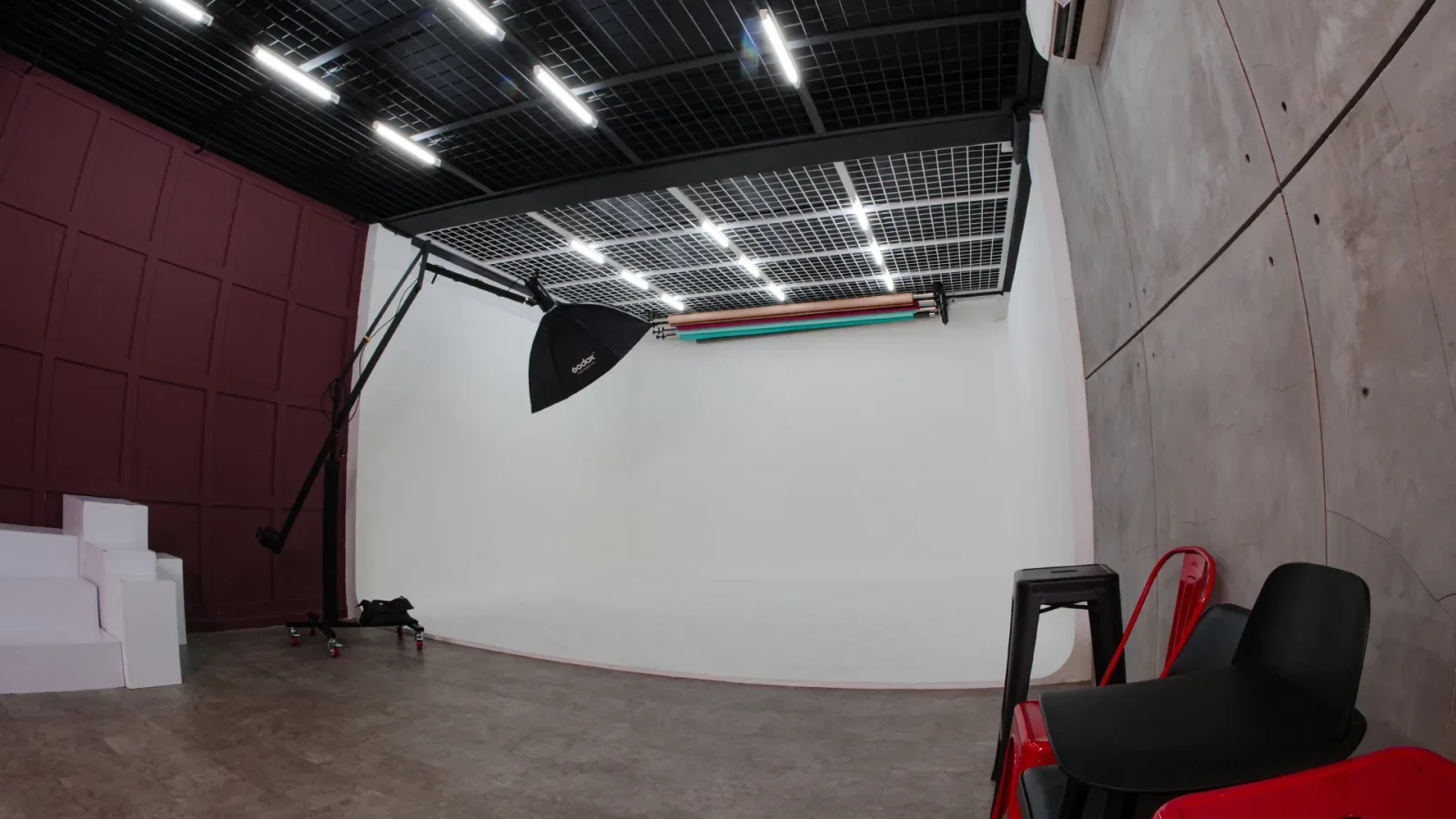 Studio Limbo kosong dengan cyclorama putih melengkung, dinding panel maroon di kiri, octabox Godox di boom stand, dan dua tabung lampu warna hijau serta merah.