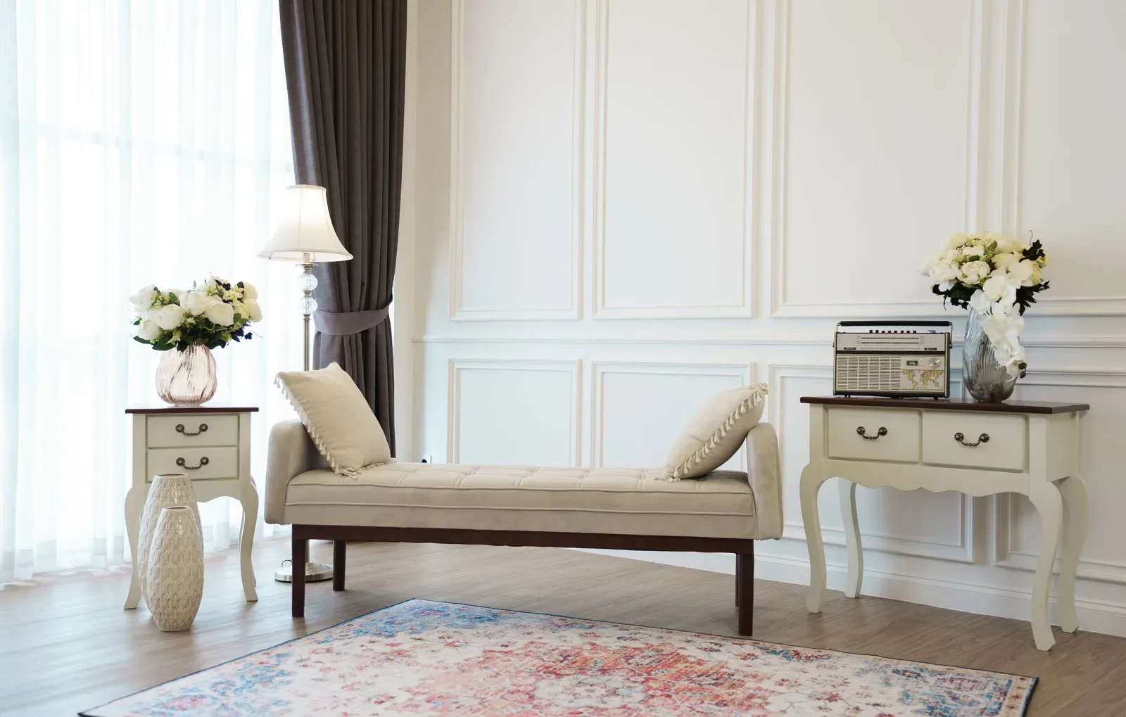 Set studio Family bergaya klasik: dinding putih berpanel moulding, daybed beige berkancing, dua konsol putih, standing lamp, dan karpet Persia pastel di lantai kayu.
