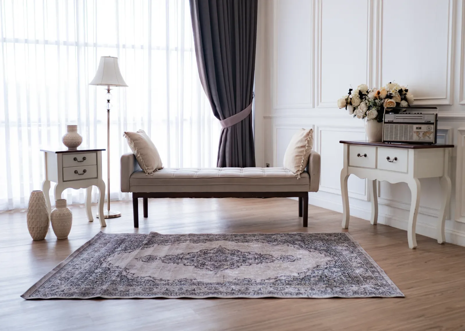 Set studio klasik Eropa serba putih dengan wainscot, tirai tembus cahaya, daybed berlapis kain krem, dua nakas berukir, radio antik, dan karpet Persia abu.