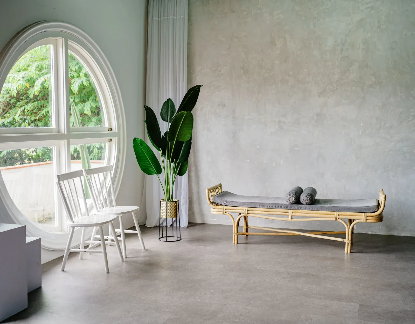 Studio Libra terang dengan jendela bulat besar bertirai sheer, dinding plester abu, daybed rotan berbantal guling, dua kursi kayu putih, dan tanaman pisang hias.