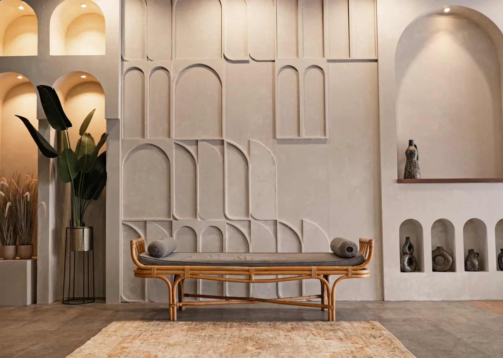 Set studio bergaya Mediterania dengan dinding plester krem berelief lengkung, ceruk berisi vas keramik, daybed rotan berbantal guling, dan tanaman pisang hias.