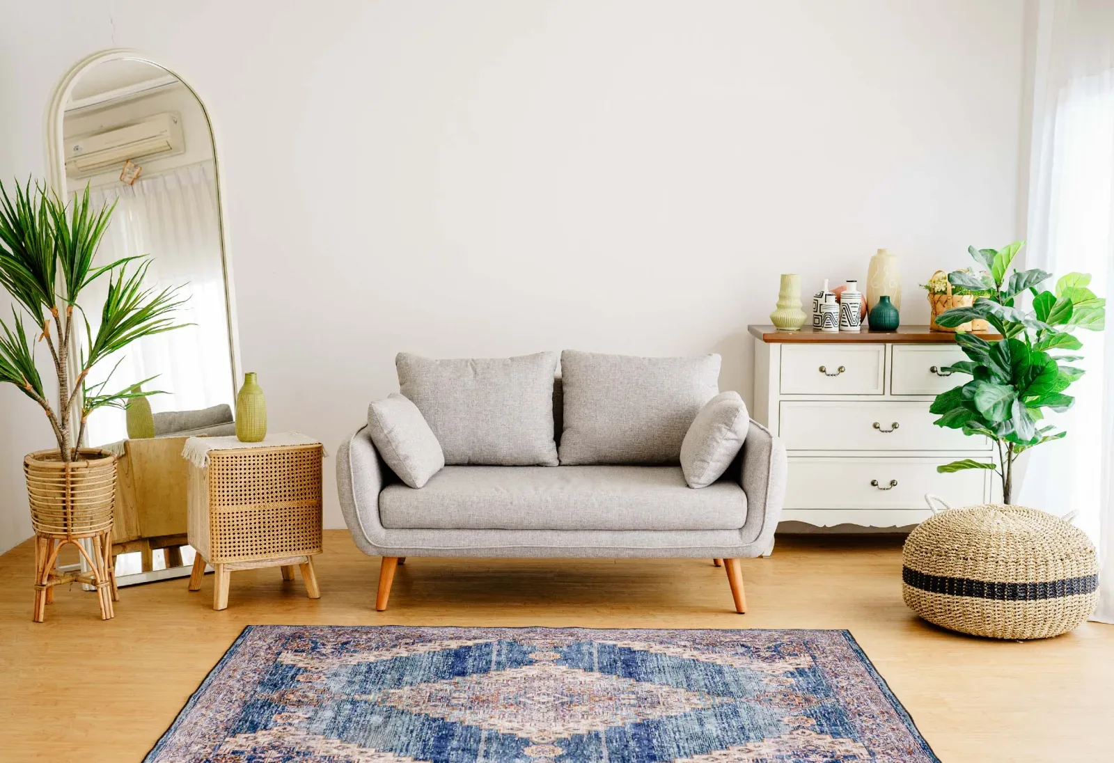 Set studio Scorpio bergaya ruang tamu: sofa abu dua dudukan, lemari laci putih bertopping kayu, cermin lengkung besar, tanaman hias, dan karpet Persia biru.