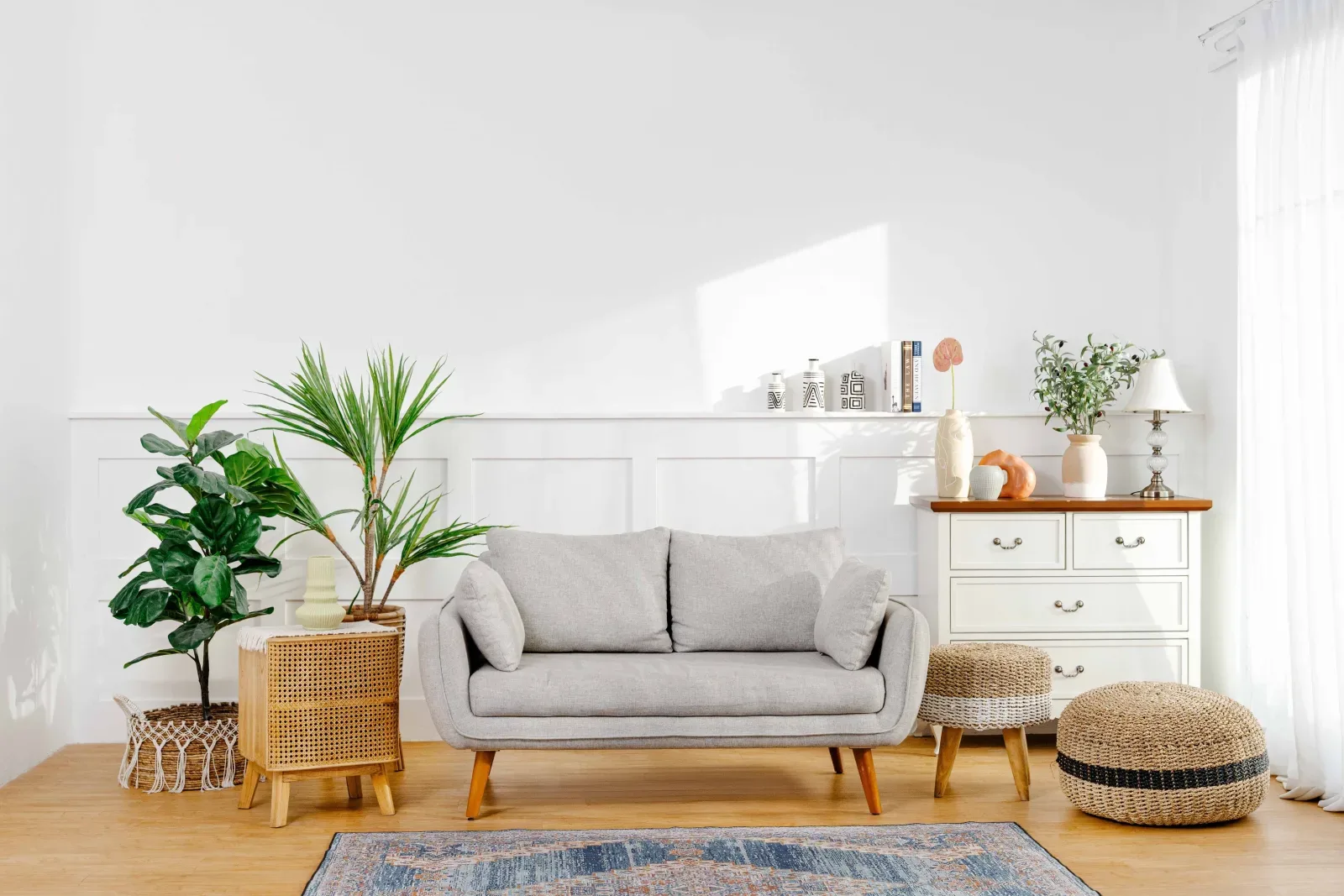 Set Scorpio dilihat lebih lebar: dinding putih berpanel wainscoting kena cahaya jendela, sofa abu, lemari laci putih berlampu meja, tanaman fiddle leaf, dan karpet Persia biru.