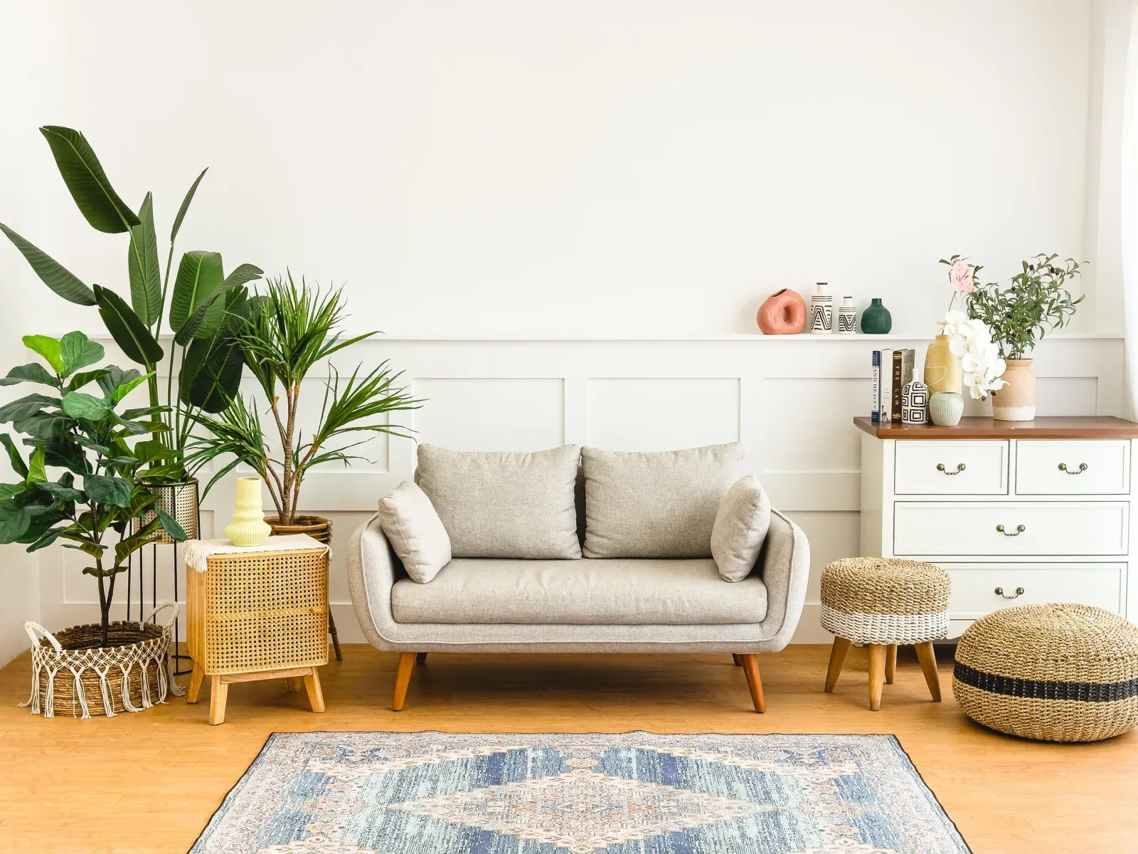 Set ruang tamu dengan sofa kain krem di depan dinding wainscoting putih, kelompok tanaman hijau besar, bufet laci putih, dua puff anyaman, dan karpet motif biru.