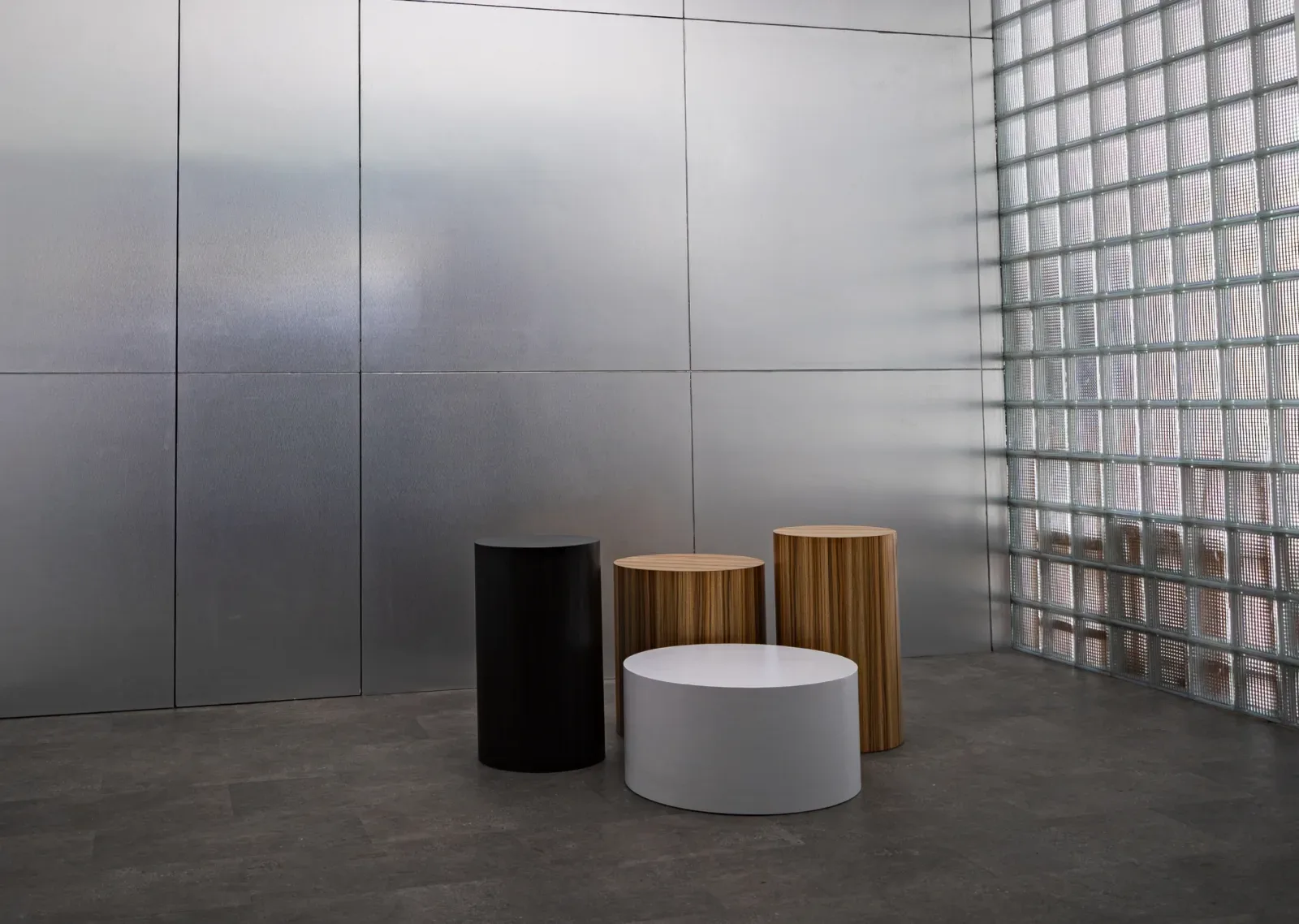 Sudut studio industrial dengan dinding panel aluminium dan dinding glass block, berisi empat podium silinder hitam, kayu, dan putih di atas lantai beton.