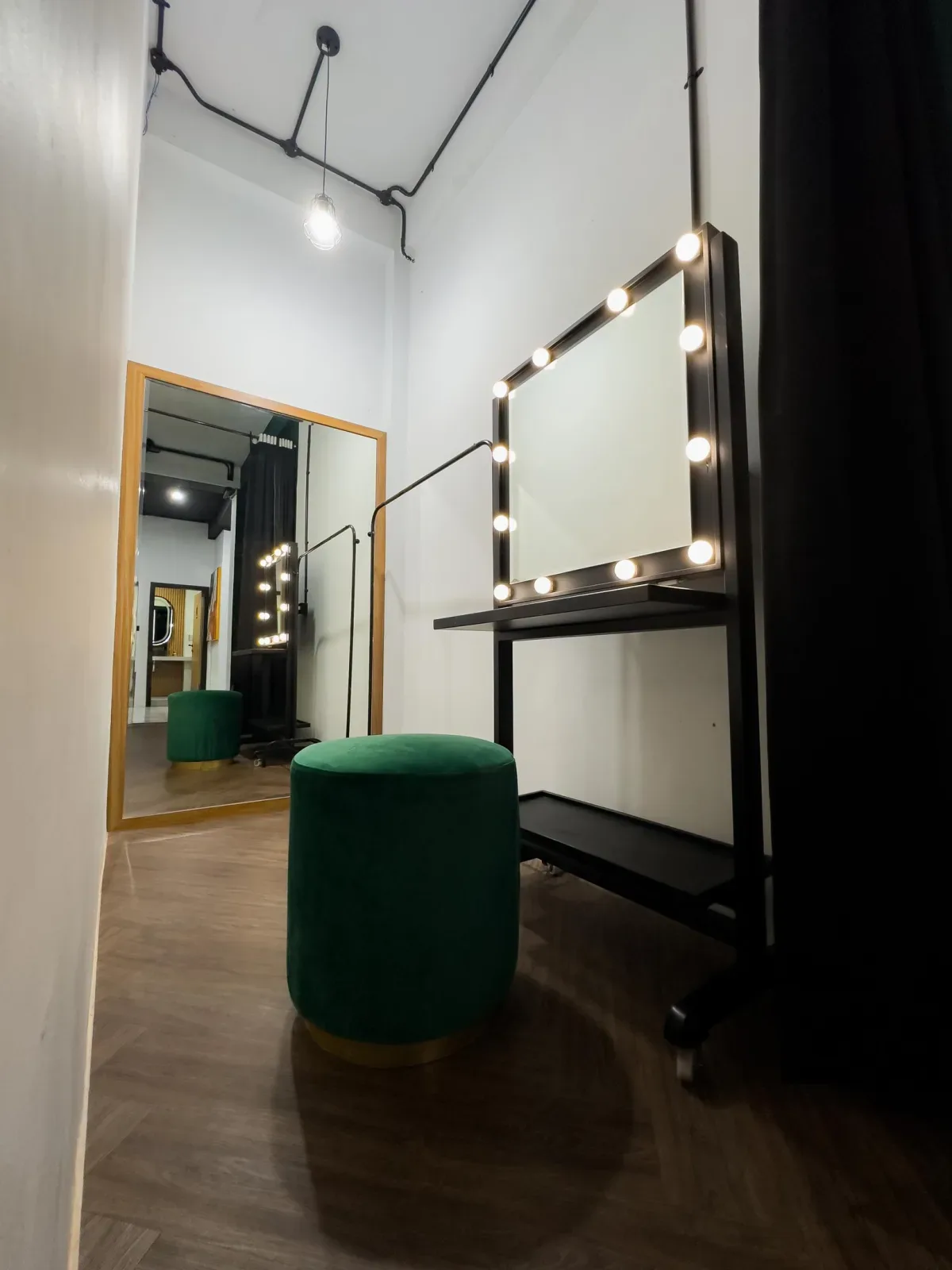 Sudut rias bergaya industrial dengan meja rias beroda rangka besi hitam, cermin persegi berlampu bohlam, puff beludru hijau, dan lantai kayu herringbone.