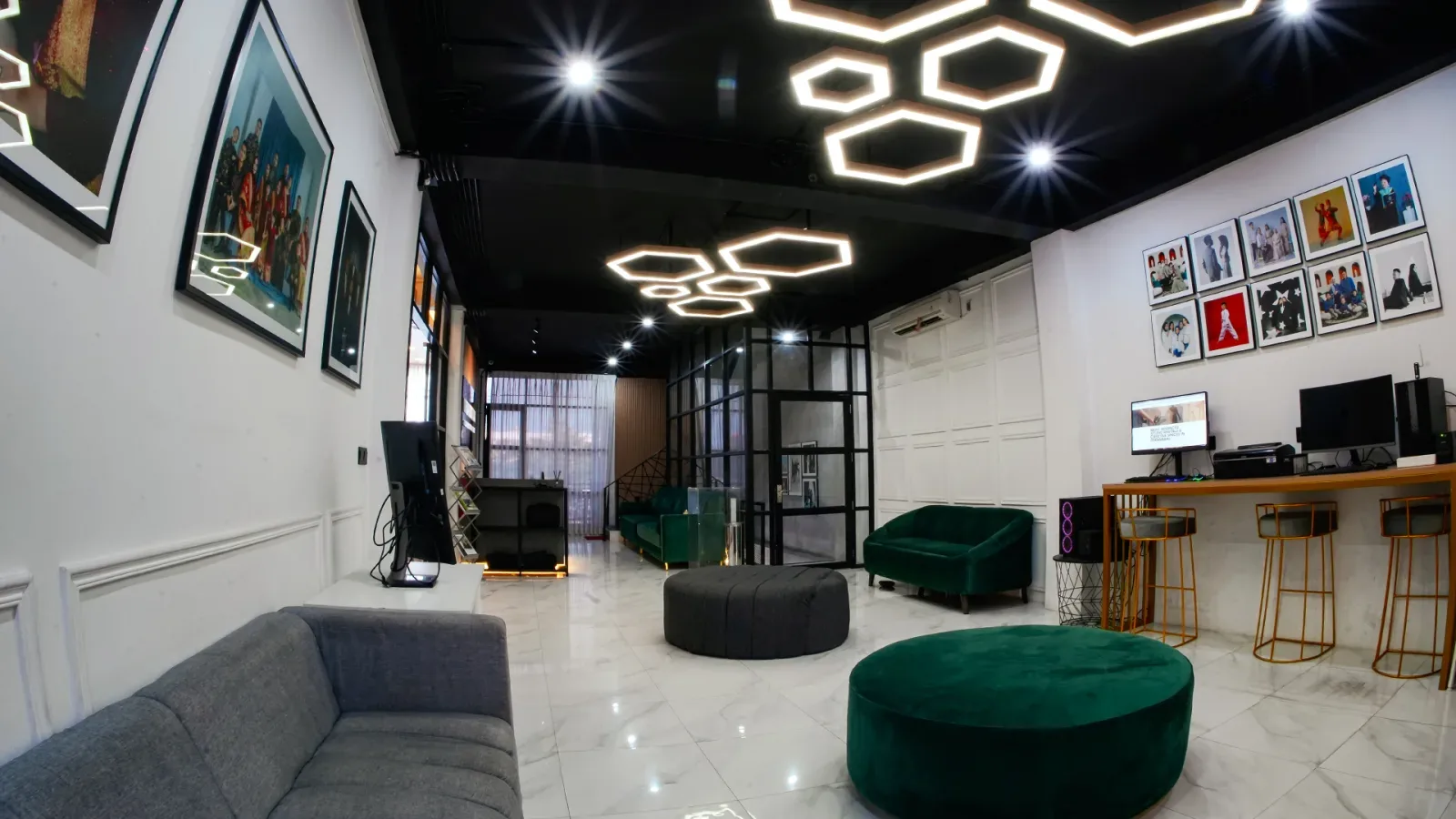 Lobi berlantai marmer putih dengan lampu gantung heksagon, sofa abu panjang, puf bundar hijau, dinding galeri berbingkai foto, dan meja bar tinggi berkursi emas.