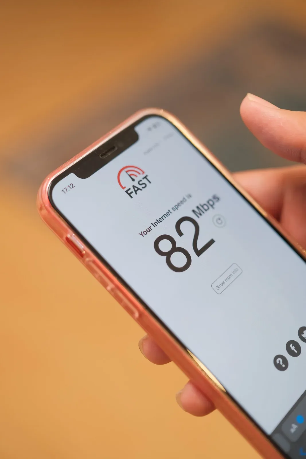 Close-up tangan memegang ponsel bercasing oranye yang menampilkan hasil tes kecepatan internet 82 Mbps dengan latar bokeh hangat.