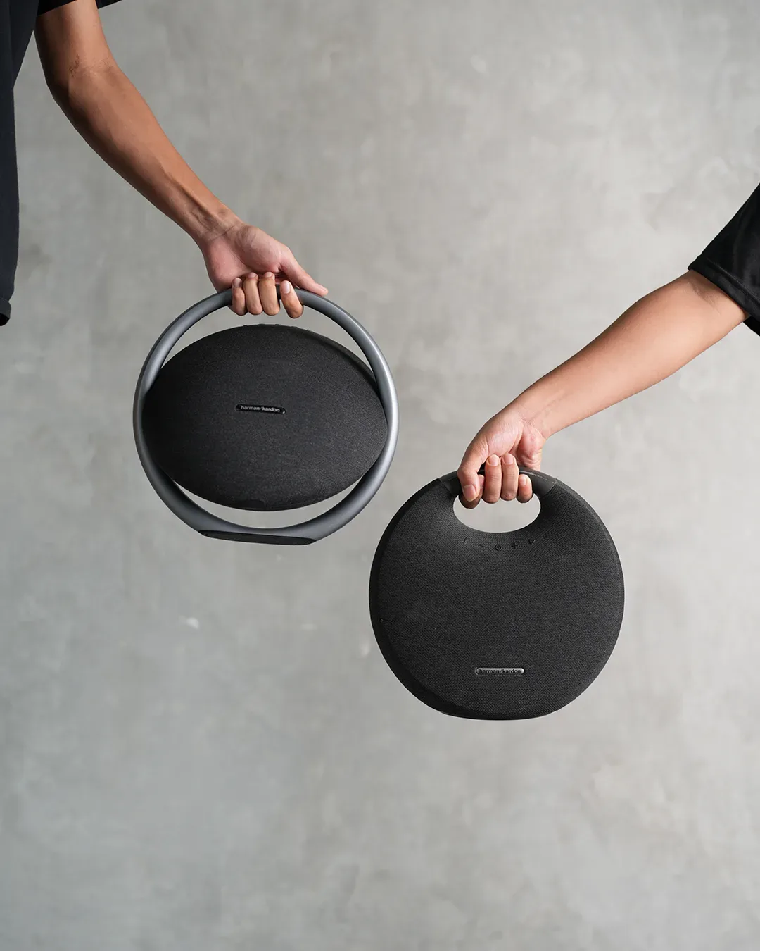 Dua tangan mengangkat sepasang speaker Bluetooth Harman Kardon Onyx Studio di depan dinding semen abu-abu.