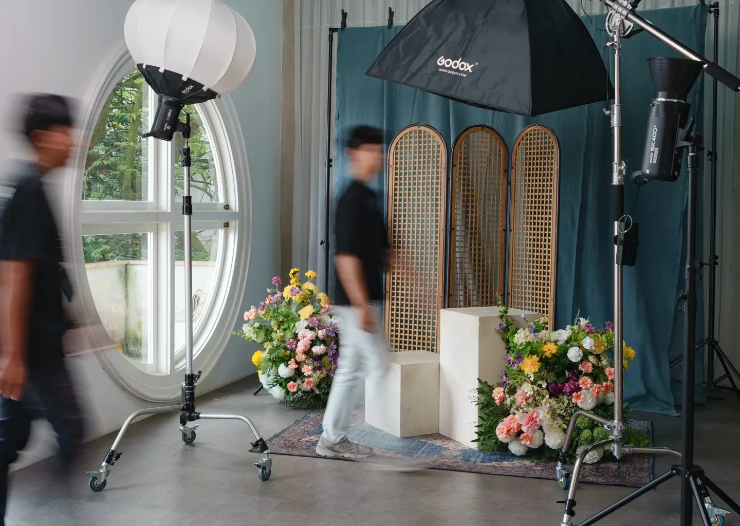Behind the scene sesi pemotretan dengan backdrop kain hijau teal, sekat lipat rotan, plinth putih, dua rangkaian bunga besar, dan dua kru berjalan blur di antara lampu.