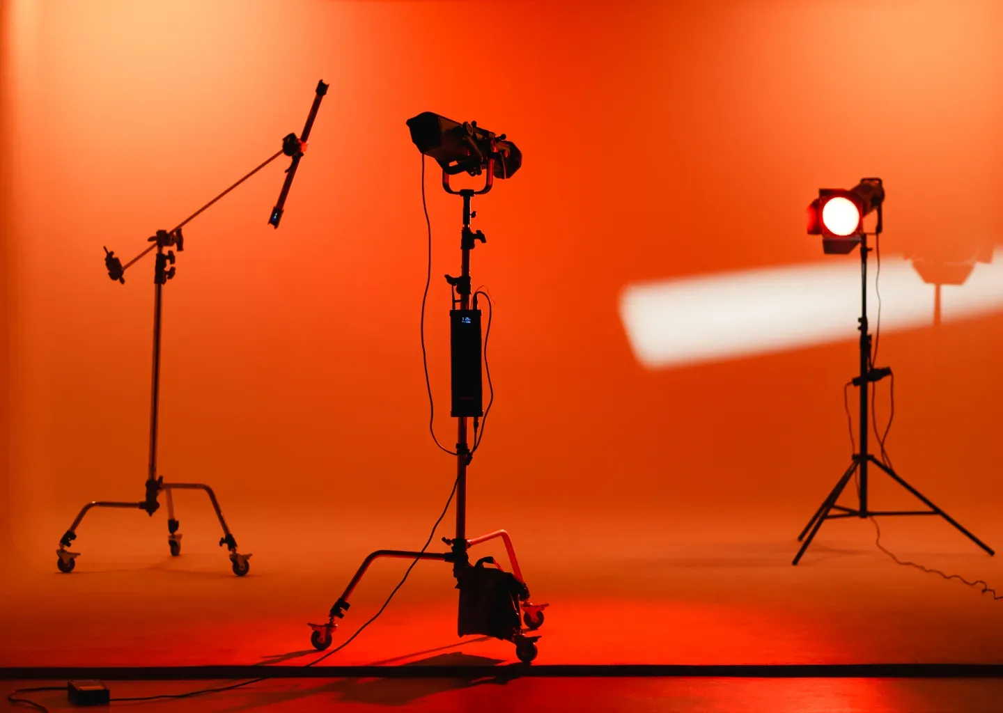 Studio kosong bermandi cahaya oranye merah dengan tiga rig lighting bersiluet: C-stand boom beroda, fresnel di stand berbaterai, dan tube light menyilang.