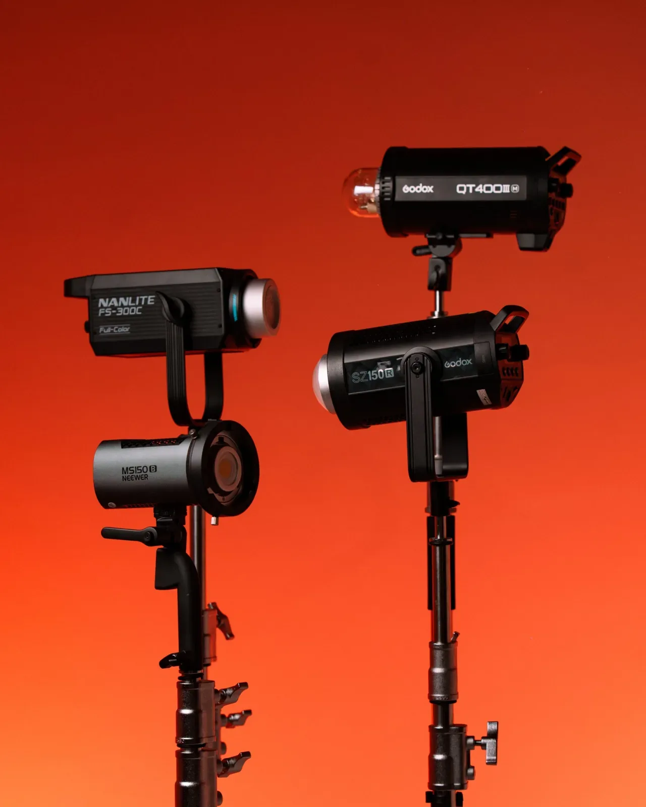 Empat unit lighting studio terpasang di light stand dengan latar gradasi oranye merah: Nanlite FS-300C, Neewer MS150B, Godox QT400 III M, dan Godox SZ150R.