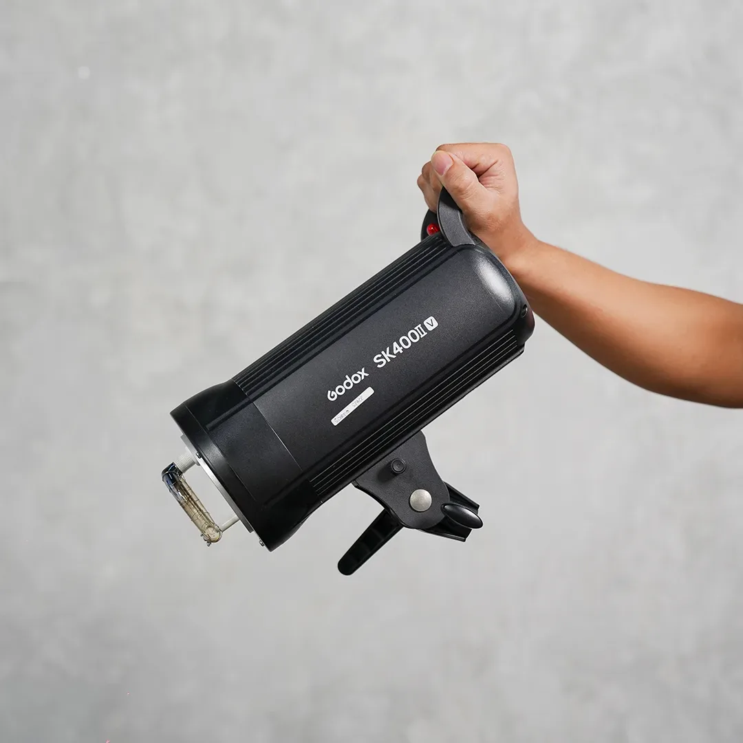 Tangan memegang lampu flash studio Godox SK400II V berbadan hitam dengan tabung flash dan modeling lamp di depan dinding beton abu.