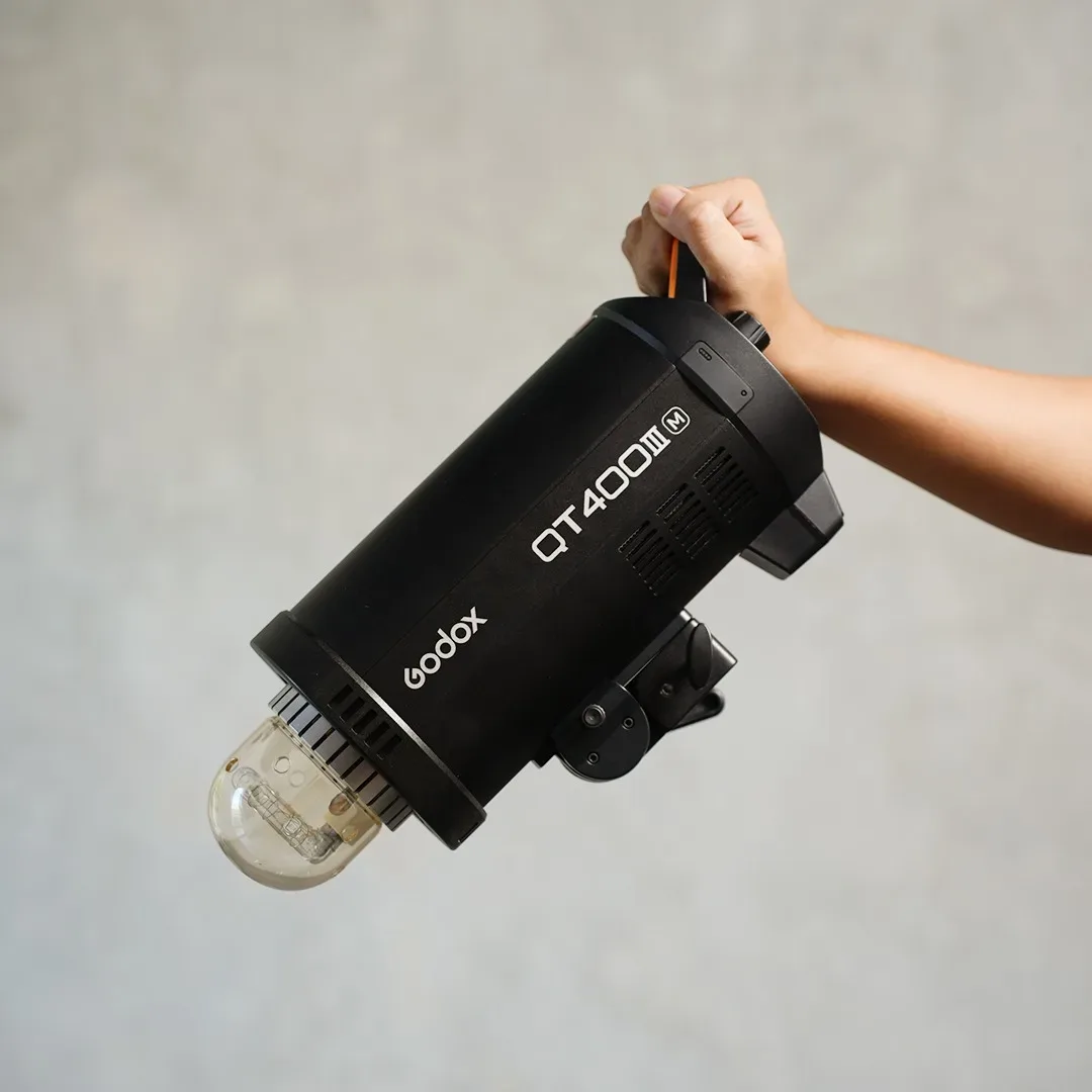 Tangan memegang lampu studio monolight Godox QT400III M berbadan hitam dengan bohlam flash terbuka di depan dinding semen abu.