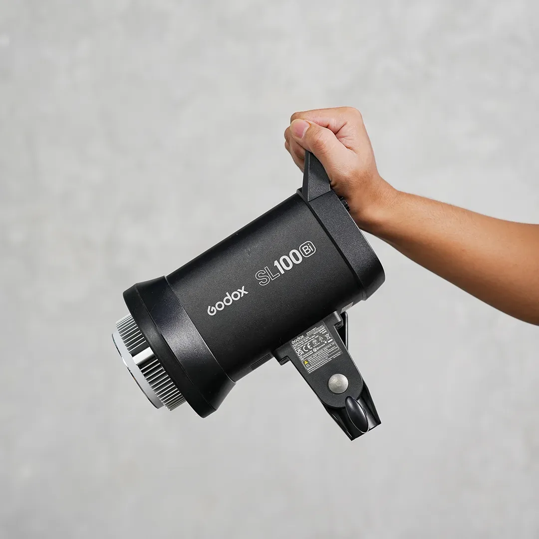 Tangan mengangkat lampu LED continuous Godox SL100Bi bi-color lewat gagangnya dengan mount Bowens terlihat di ujung depan.