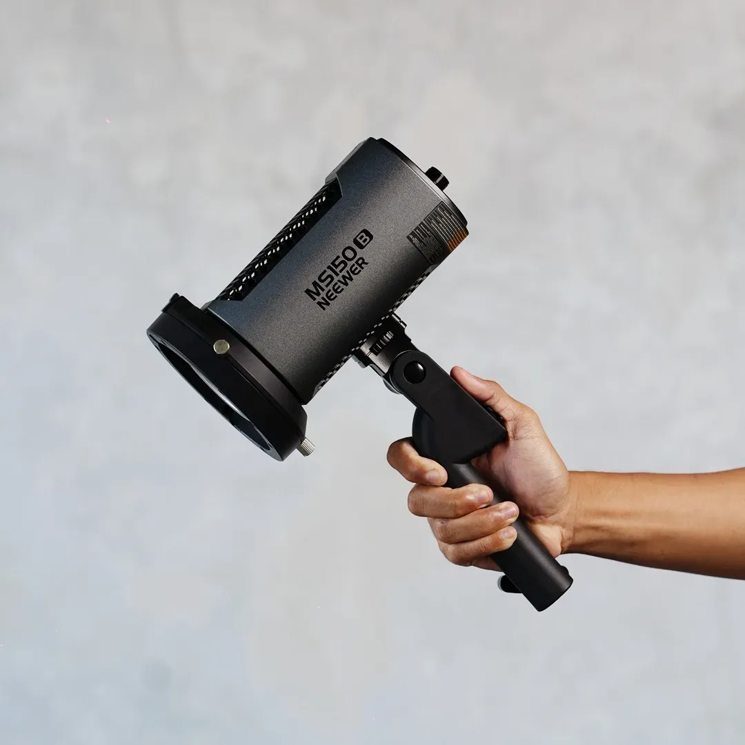 Lampu LED monolight Neewer MS150 B abu gelap dipegang lewat grip handle hitam di depan dinding plester abu.
