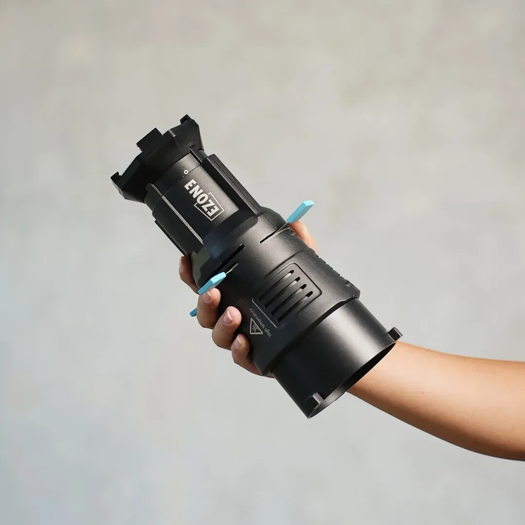 Tangan memegang optical spotlight silinder hitam bermerek Ezone dengan tuas pengatur biru muda dan lubang ventilasi.
