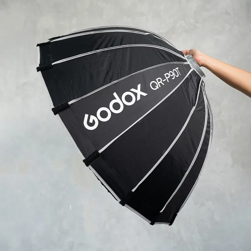 Softbox parabolik lipat hitam berusuk perak bertuliskan Godox QR-P90T dipegang tangan di depan dinding semen abu.