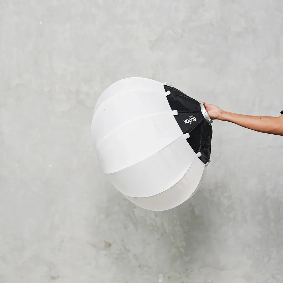 Softbox lantern bulat Godox CS65 berkain putih dengan skirt hitam dipegang satu tangan di depan dinding beton abu.