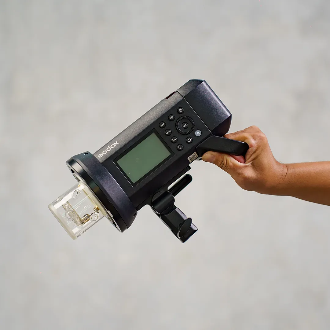 Lampu flash monolight bertenaga baterai Godox dipegang tangan, terlihat layar LCD, roda kontrol putar, dan tabung bare-bulb berpelindung kaca.