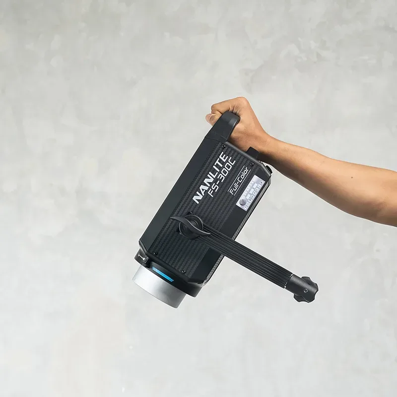 Tangan memegang lampu LED monolight Nanlite FS-300C Full-Color pada gagangnya dengan yoke terpasang di depan dinding semen abu.