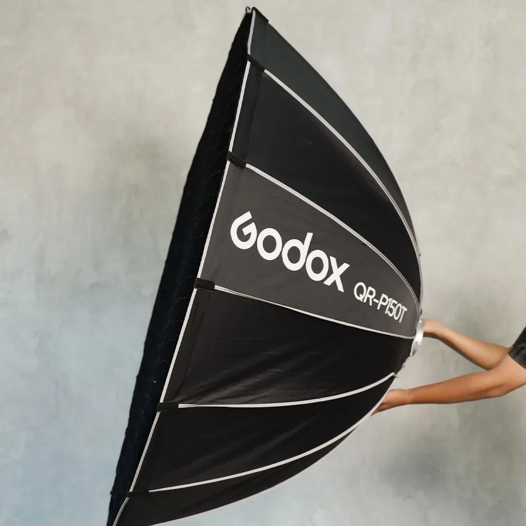 Tangan memegang softbox parabolik lipat hitam besar bertuliskan Godox QR-P150T di depan dinding semen abu bertekstur.