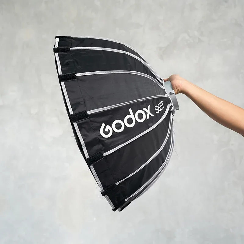 Softbox lipat hitam berukuran kecil dengan rusuk perak bertuliskan Godox S65T dipegang tangan di depan dinding semen abu.