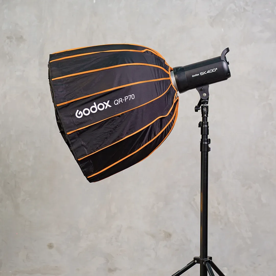 Lampu studio Godox SK400 II di light stand hitam memakai softbox parabolik Godox QR-P70 hitam berusuk oranye.