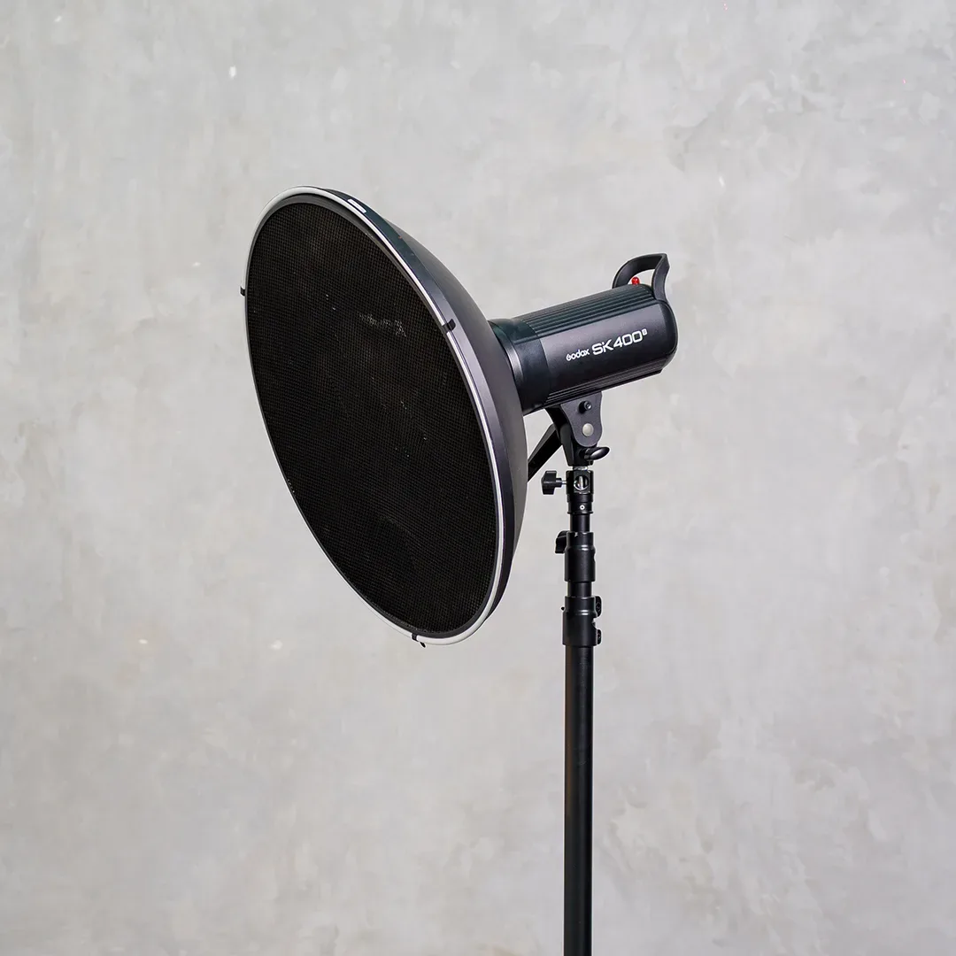 Lampu studio Godox SK400II di light stand memakai beauty dish bergrid honeycomb hitam, difoto dari samping di depan dinding beton abu.
