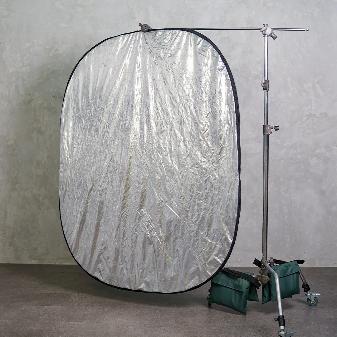 Reflektor lipat oval besar sisi silver disandarkan pada boom arm C-stand beroda dan ditahan dua sandbag hijau di ruang berlantai semen.