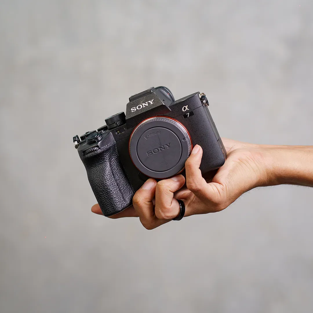 Bodi kamera mirrorless full-frame Sony Alpha a7 dengan tutup bodi E-mount terpasang digenggam satu tangan di depan latar abu blur.