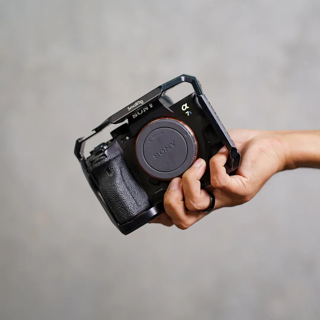 Tangan memegang bodi kamera Sony Alpha a7S III tanpa lensa yang dibungkus cage SmallRig di depan dinding semen abu.
