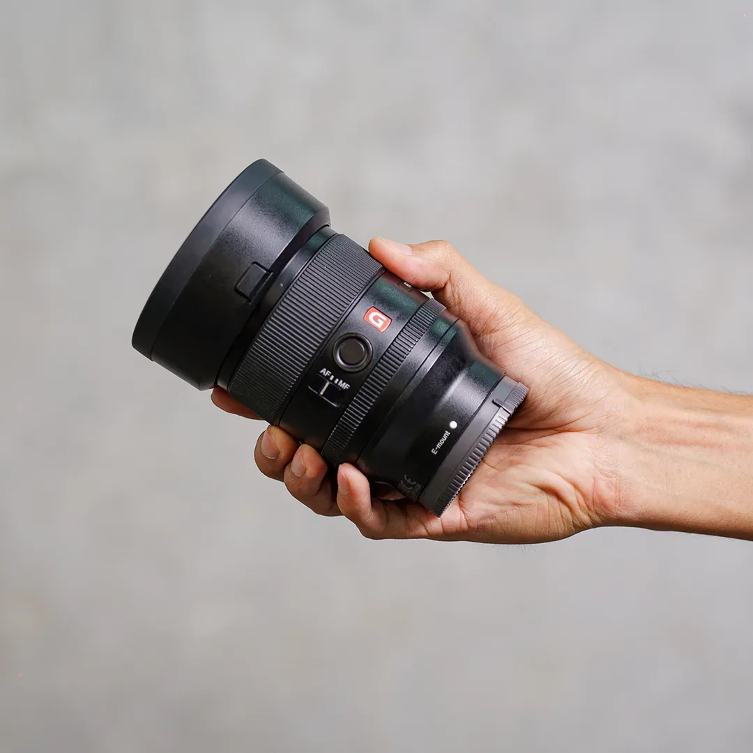 Tangan memegang lensa prime Sony E-mount seri G Master dengan badge merah, sakelar AF/MF, dan hood bunga terpasang.