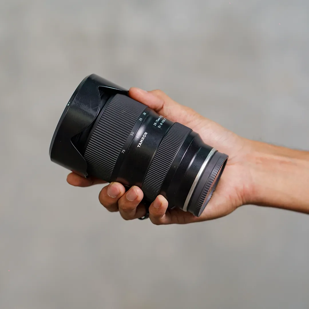 Tangan memegang lensa zoom Tamron 28-75mm f/2.8 dengan lens hood terpasang, latar dinding semen abu blur.