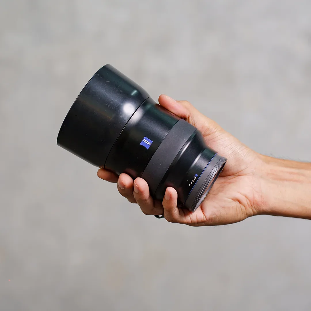 Tangan memegang lensa Zeiss E-mount berbadan hitam dengan badge biru ZEISS dan hood terpasang di depan dinding semen abu.