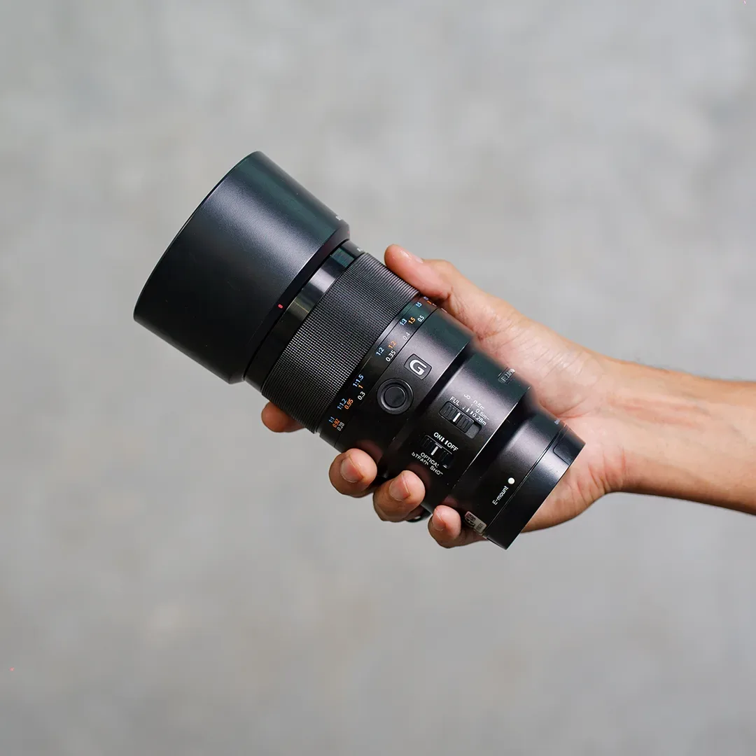 Lensa makro Sony G bertudung besar digenggam tangan; terlihat badge G, tombol focus hold, saklar limiter fokus, dan saklar SteadyShot.