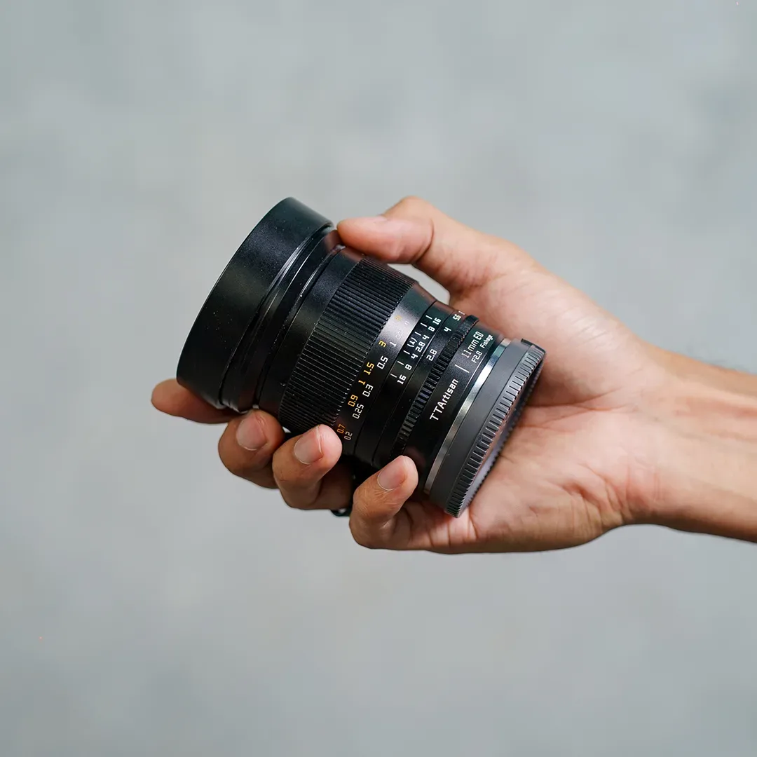 Lensa fisheye TTArtisan 11mm f/2.8 ED digenggam telapak tangan dengan hood logam menyatu terlihat jelas.