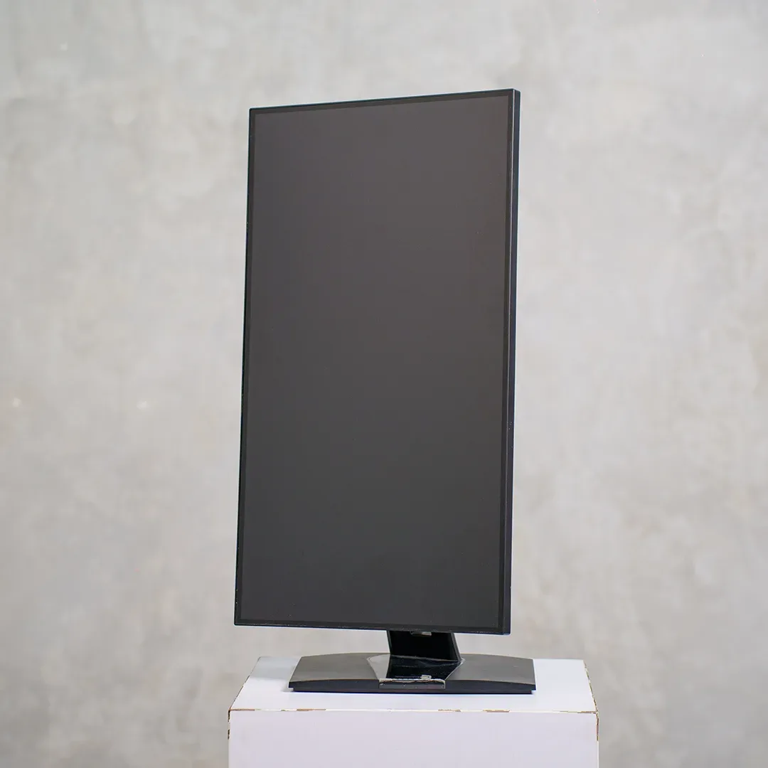 Monitor layar datar bezel tipis dipasang vertikal di atas dudukan hitam mengilap, diletakkan di atas kotak putih dengan latar dinding beton abu.