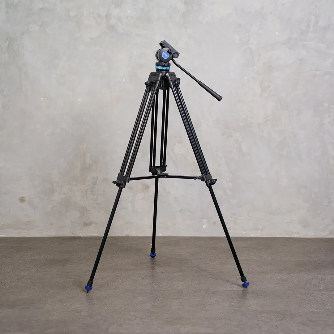 Tripod video hitam berkaki tiga dengan fluid head dan tuas pan beraksen biru berdiri di lantai semen di depan dinding beton abu.