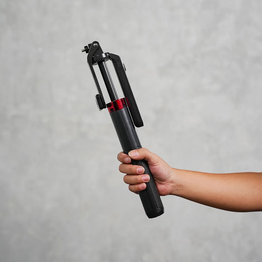 Tangan memegang tripod ponsel mini lipat hitam beraksen merah lengkap dengan penjepit smartphone dan ball head kecil.