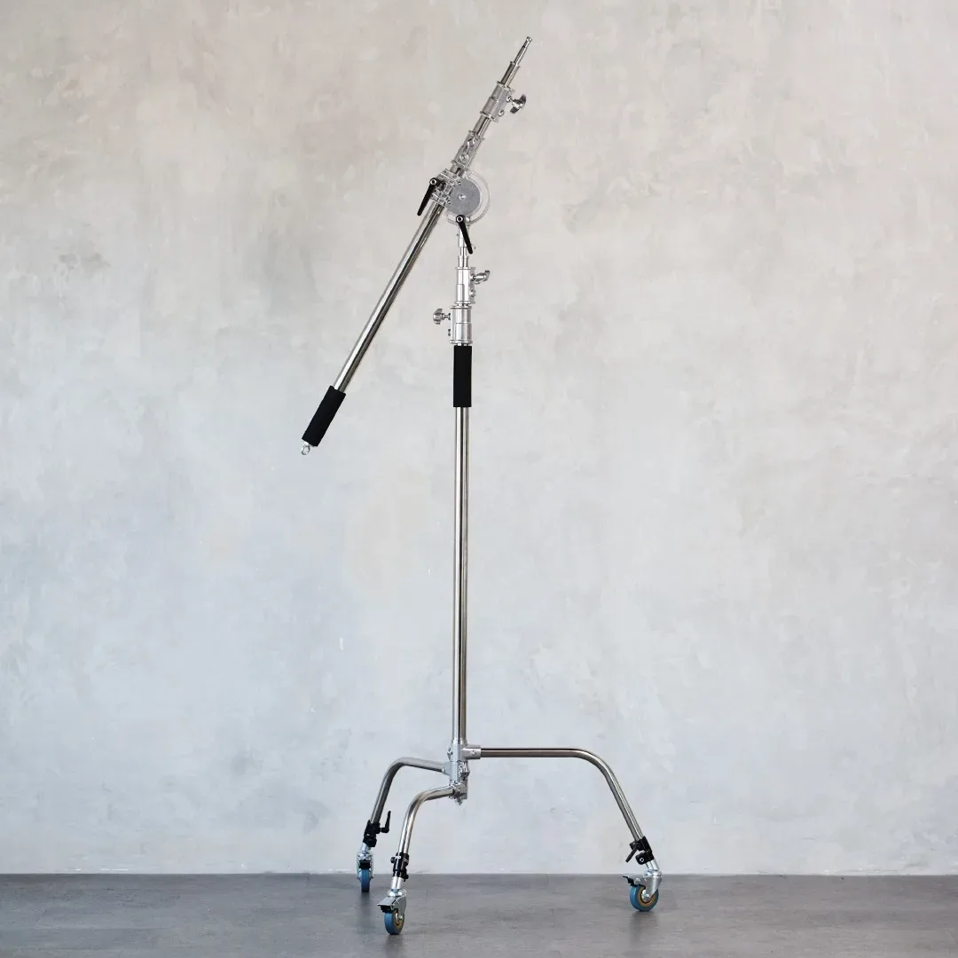 C-stand krom berat dengan boom arm terpasang miring dan kaki beroda berdiri di depan dinding plester abu.