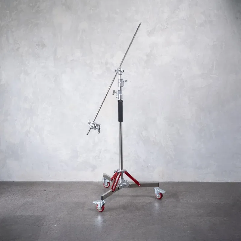 Light stand heavy duty beroda dengan lengan boom panjang terpasang miring, tiang krom dan penopang merah, di depan dinding semen abu.