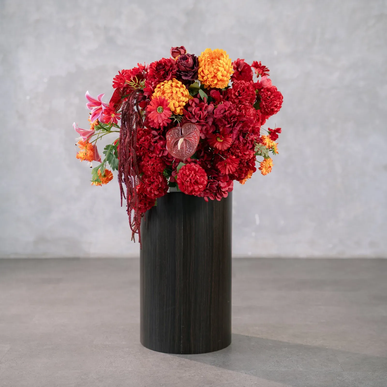 Rangkaian bunga artifisial merah oranye dengan amaranthus menjuntai di dalam vas silinder kayu gelap, berdiri di lantai beton.