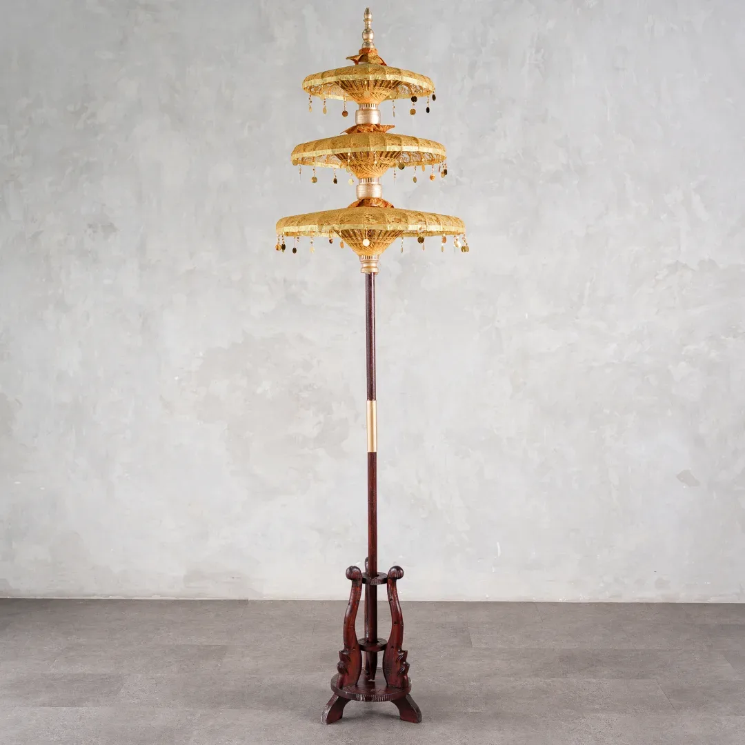 Payung susun tiga tingkat warna emas bergaya Bali berumbai koin logam dipasang pada tiang kayu berkaki tripod jati berukir.