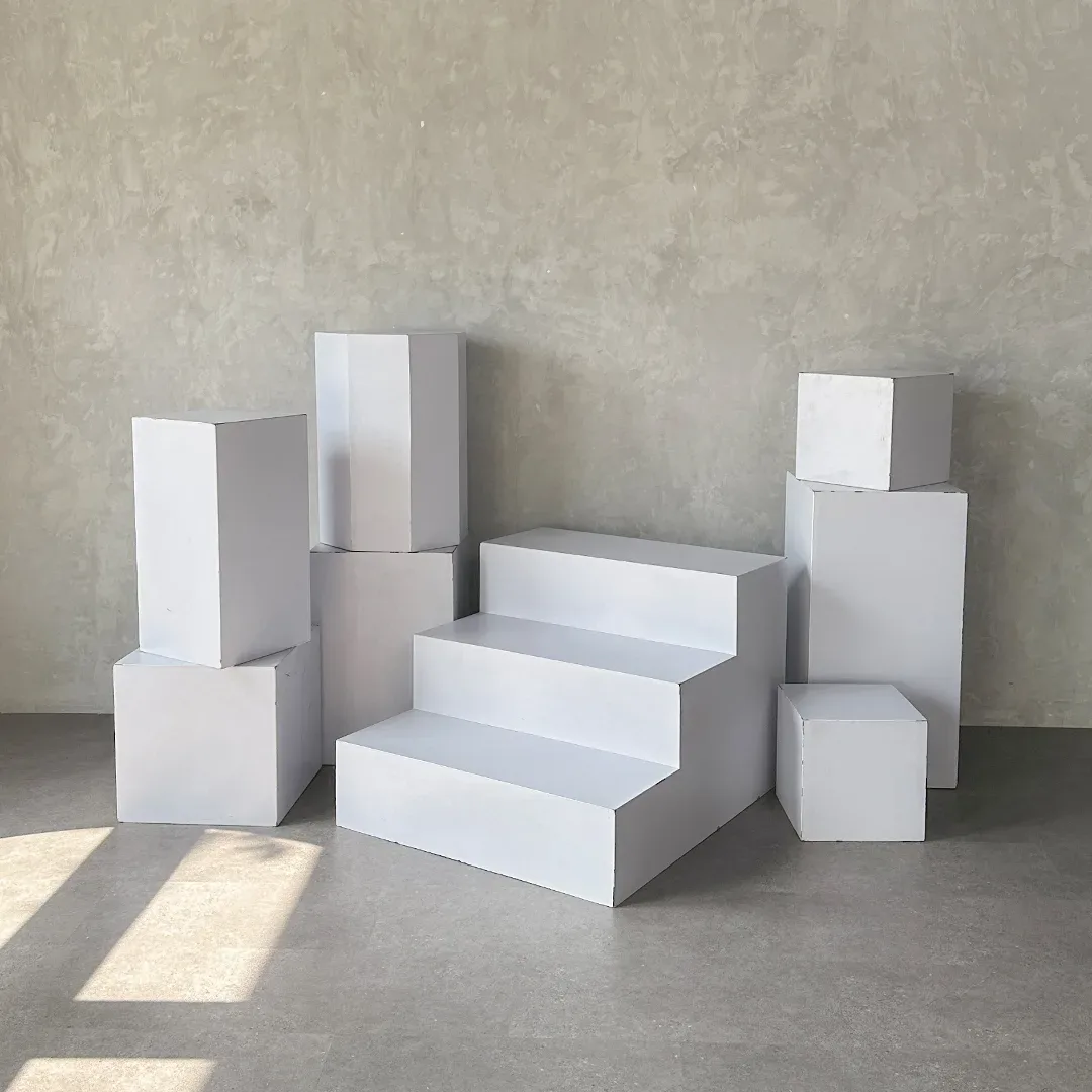 Susunan kubus podium putih berbagai ukuran ditumpuk di lantai beton dengan satu tangga podium tiga anak tangga putih di tengah.