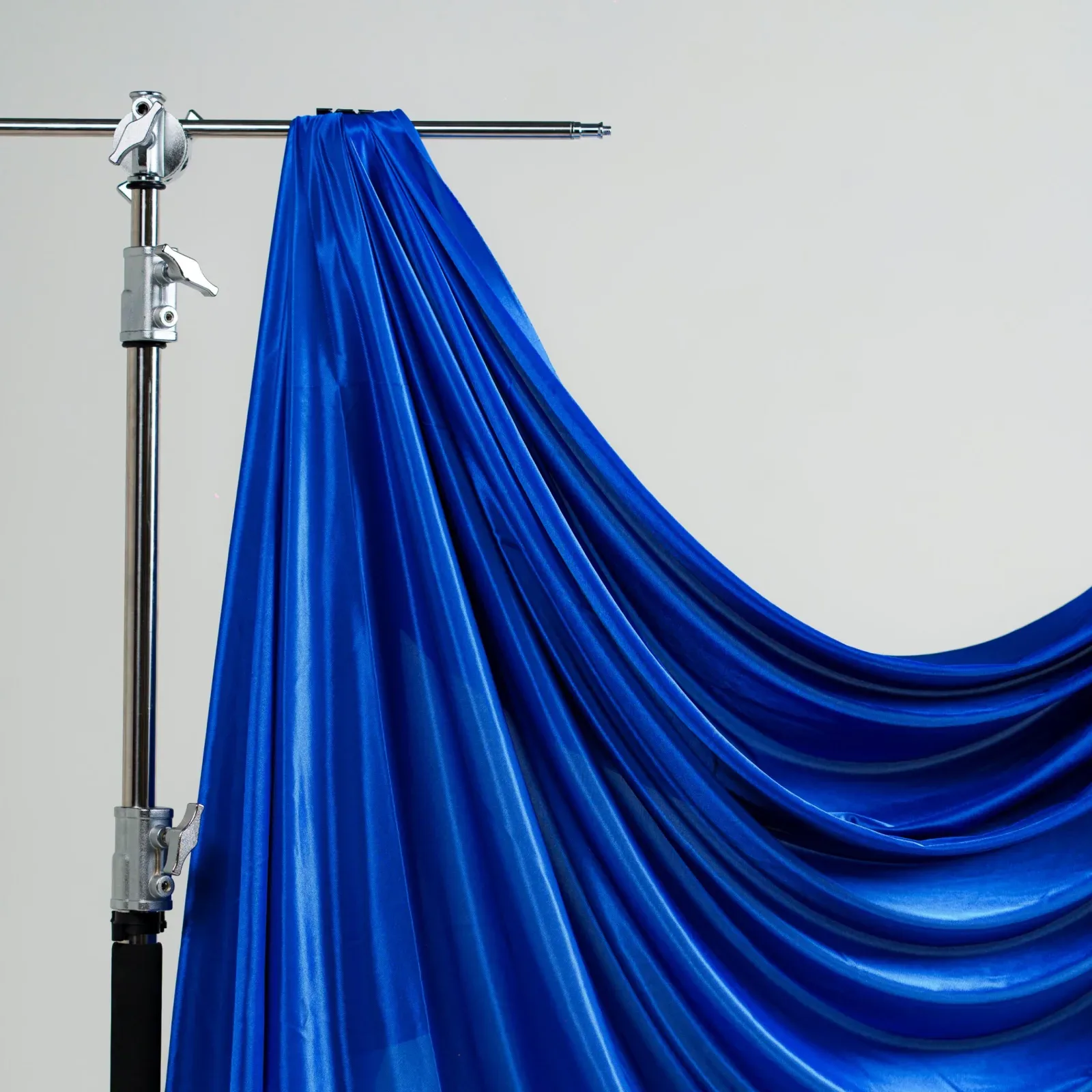 Kain latar biru royal berbahan satin mengkilap digantung di crossbar C-stand dan jatuh berlipat dengan latar dinding abu polos.