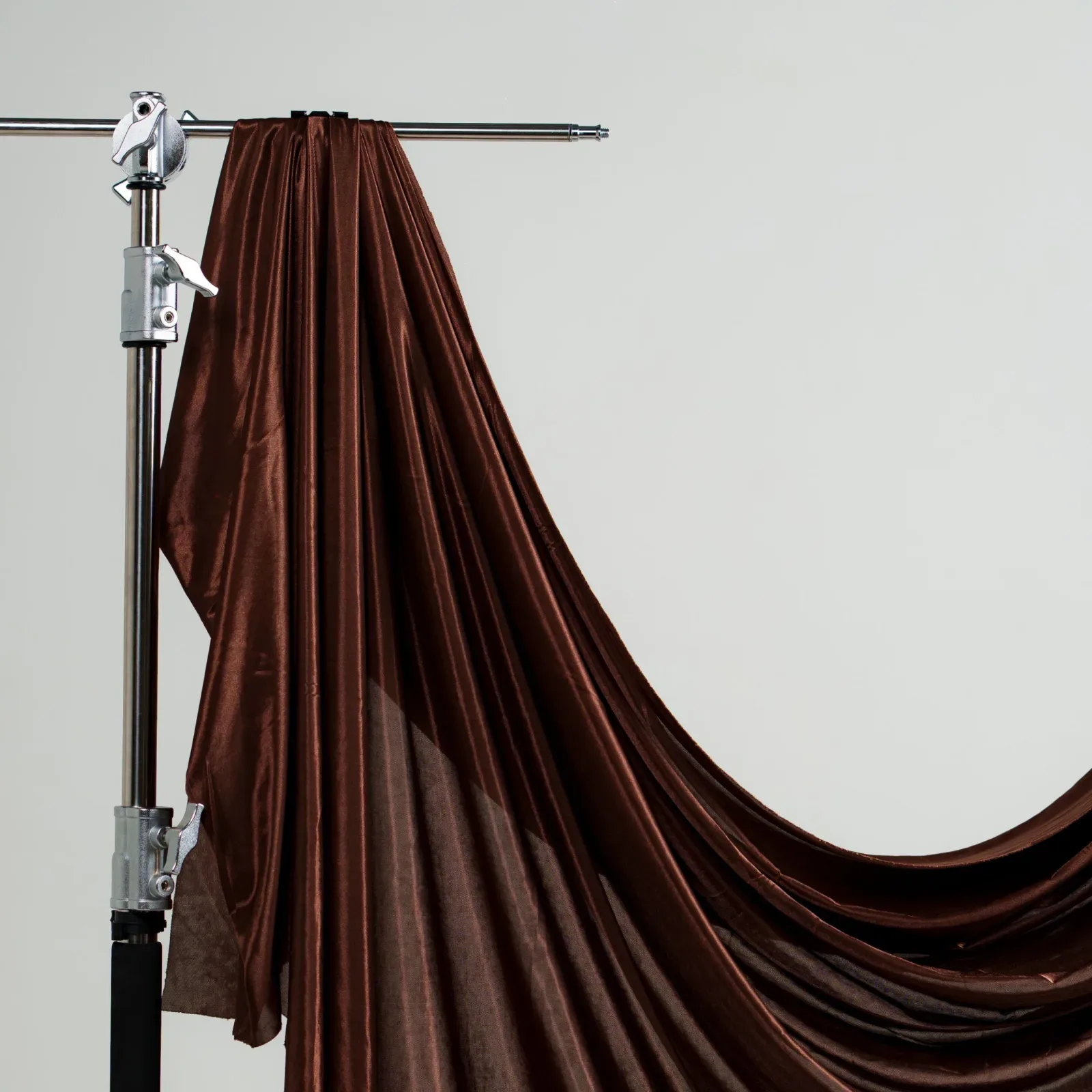 Kain latar satin coklat mengilap digantung dari boom arm C-stand krom dan jatuh berlipat dengan latar dinding studio abu polos.