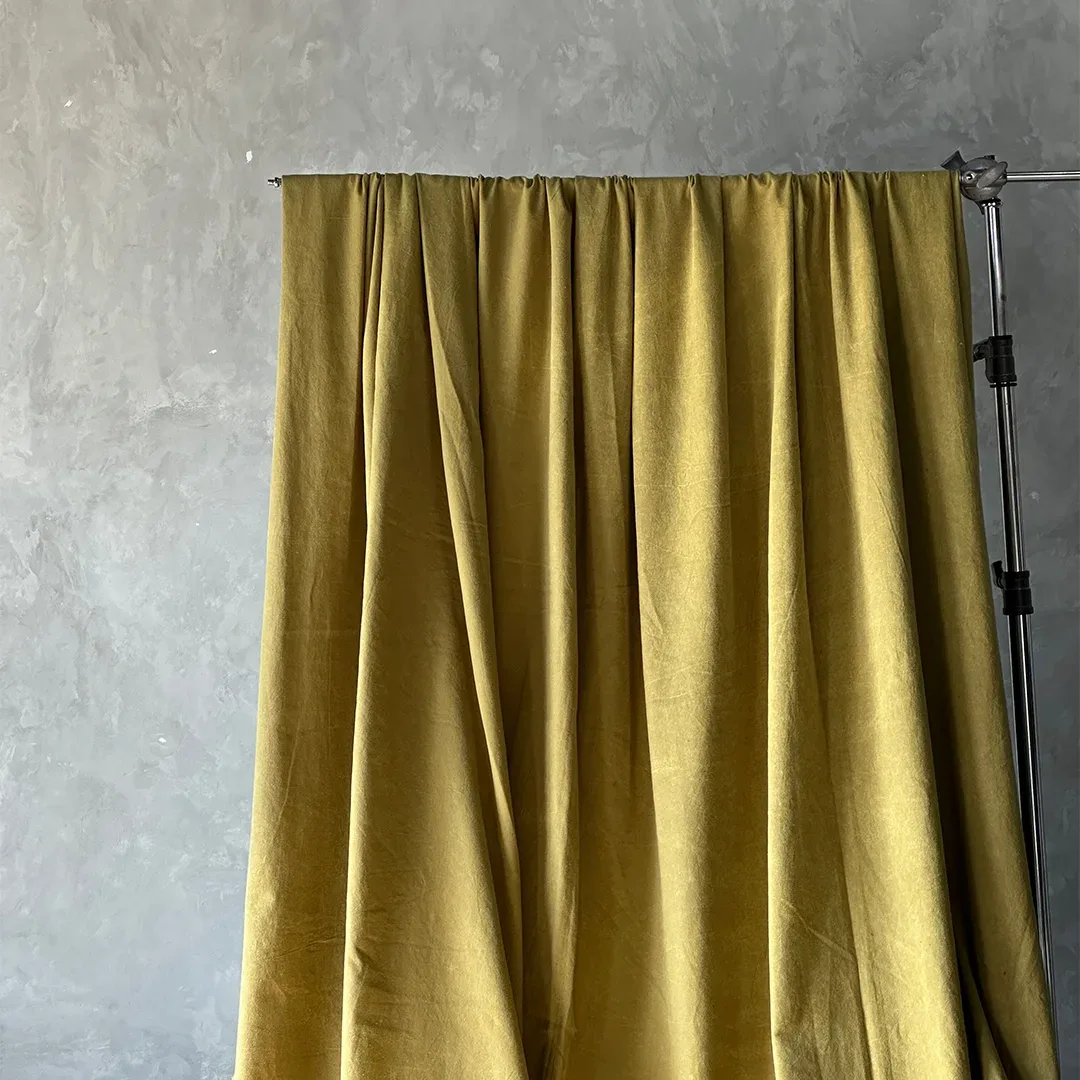 Kain latar polos warna mustard kehijauan tergantung di batang besi bertopang C-stand dan jatuh berlipat ke lantai.