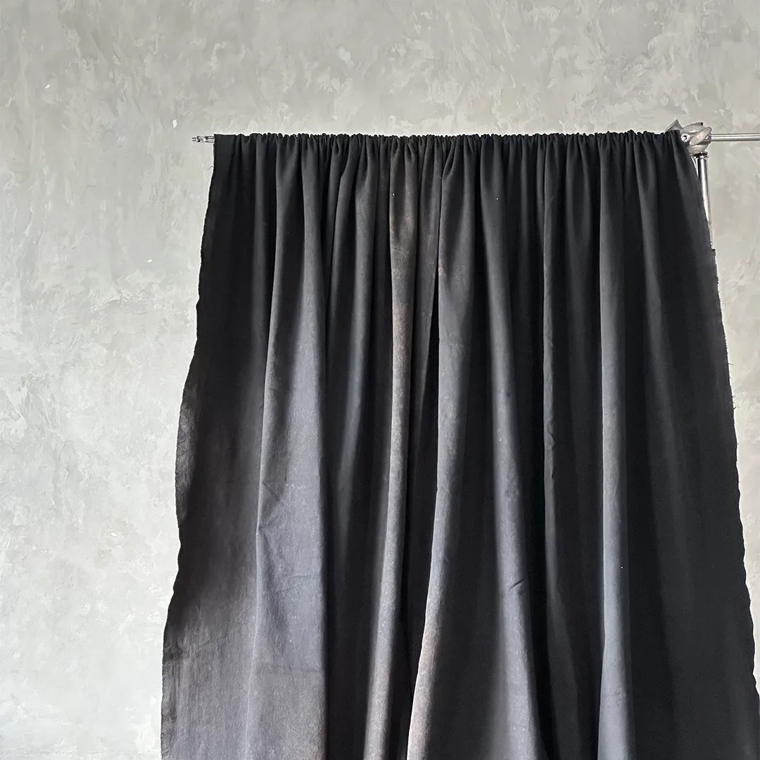 Kain latar hitam digantung memanjang pada batang besi yang dijepit ke light stand, berlipat vertikal di depan dinding plester abu.