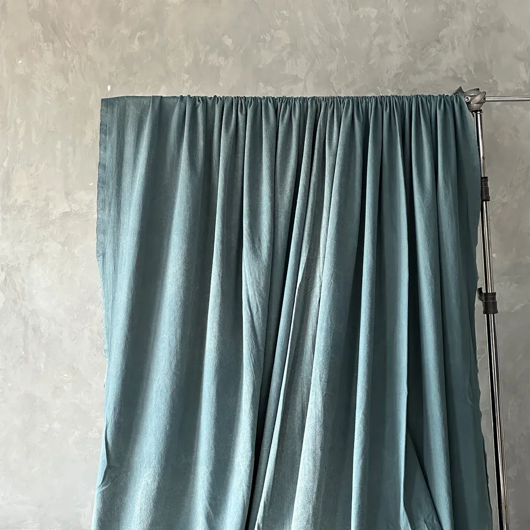 Kain latar biru kehijauan bertekstur katun digantung pada batang yang dijepit light stand dengan lipatan lembut.